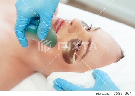 Beautiful woman having guasha scraper face massage done by cosmetologist 75834364