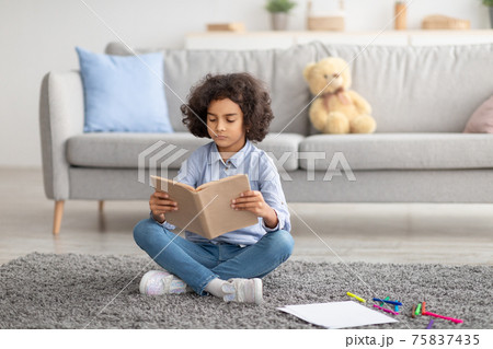 Cute black girl sitting on floor carpet, reading book 75837435