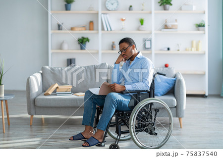 Disabled black guy in wheelchair reading notebook, working or studying from home during covid-19 epidemic 75837540