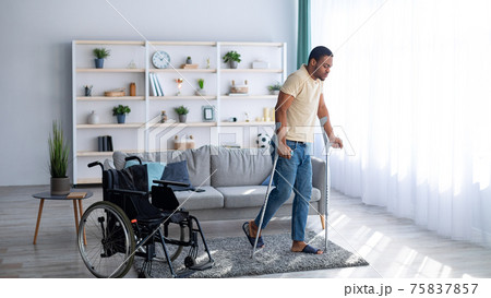 Rehabilitation concept. Young black guy walking with crutches after using wheelchair at home, panorama Rehabilitation concept. Young black guy walking with crutches after using wheelchair at home, panorama 75837857