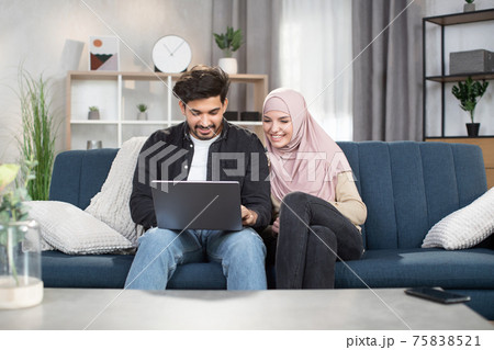 Cheerful happy Muslim couple doing online shopping in internet at home indoors, using laptop pc and sitting on comfortable blue sofa in living room. Buying goods easily, stay safe 75838521