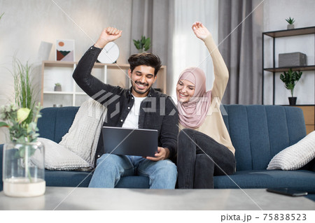 Cheerful young Muslim couple, holding hands up in victory gesture, celebrating success. Arabian couple using laptop for watching sports match together sitting on comfortable sofa in cozy living room 75838523