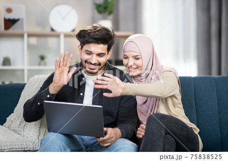 Laughing cheerful young muslim couple sitting on a couch with a laptop at home, waving while having video call talking with relatives or friends. Family, technology, wifi internet concept Laughing cheerful young muslim couple sitting on a couch with a laptop at home, waving while having video call talking with relatives or friends. Family, technology, wifi internet concept 75838525