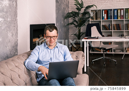 Young successful man in shirt and glasses smiling while looking into laptop while lying on sofa at home office. The concept of success, remote work and time management. Young successful man in shirt and glasses smiling while looking into laptop while lying on sofa at home office. The concept of success, remote work and time management. 75845899