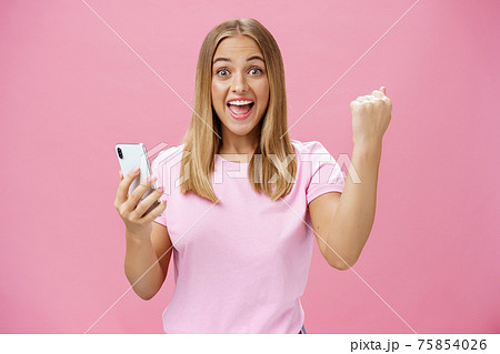 Girl beat own record in smartphone game raising clenched fist in cheer and triumph holding cellphone, smiling excited and happy at camera celebrating victory with joyful gesture over pink background 75854026