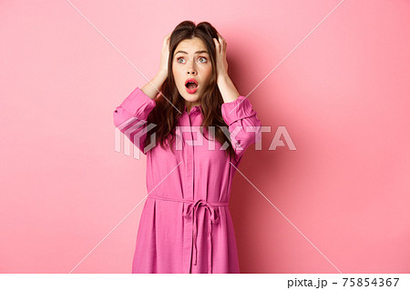 Young scared woman holding hands on head and gasping troubled, staring left at copyspace, standing over pink background 75854367
