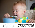 Focused adorable toddle in colorful bib sitting in highchair and eating porridge with spoon. 75857536