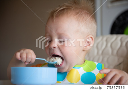 Focused adorable toddle in colorful bib sitting in highchair and eating porridge with spoon. 75857536