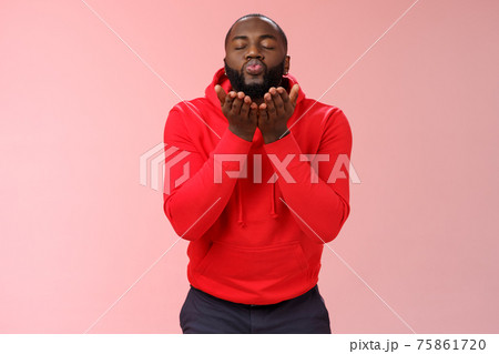 Charming cute african american bearded guy in red hoodie close eyes folding lips hold palms near mouth sending passionate air kiss blowing mwah camera, standing lovely pink background 75861720
