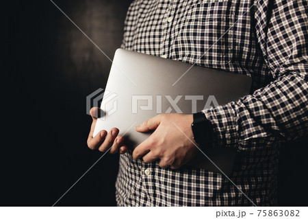 Man wearing casual clothes holds closed laptop, black blurred background 75863082