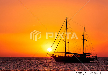 Silhouette of sailing boat with sails down against sun at sunset, sun glare on sea waters. Romantic seascape, sun touch headsail ropes. Silhouette of sailing boat with sails down against sun at sunset, sun glare on sea waters. Romantic seascape, sun touch headsail ropes. 75863136