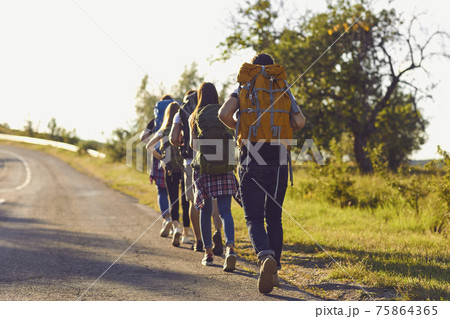 Group of young hikers walk in a row along the road with backpacks on their shoulders. Group of young hikers walk in a row along the road with backpacks on their shoulders. 75864365