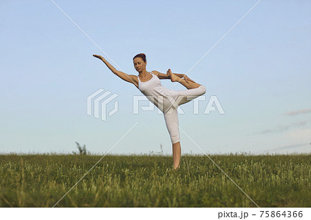 Slim yoga instructor doing Standing Bow pose on green summer meadow under clear blue sky 75864366