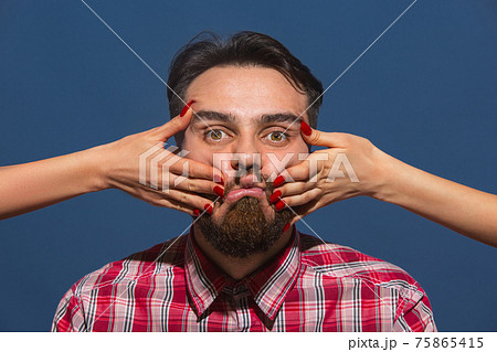 Female hands making different emotions of young bearded man on blue background Female hands making different emotions of young bearded man on blue background 75865415