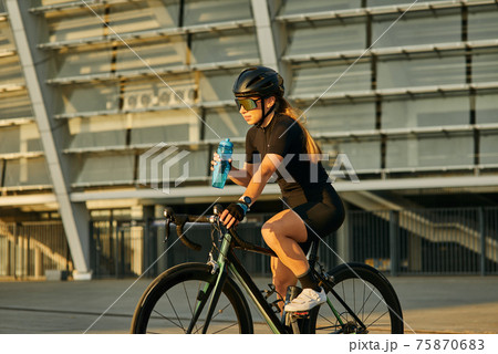 Professional female cyclist in black cycling garment and protective gear holding water bottle while riding bicycle in city, training outdoors at sunset 75870683