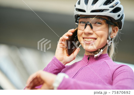 Adorable young female cyclist wearing protective helmet and glasses smiling at camera while talking on the phone, standing outdoors on a daytime 75870692