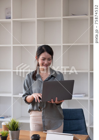Young asian woman standing and holding laptop browsing internet at home office. 75874532