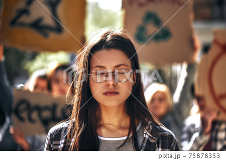 Dreaming about clean earth. Young woman in eyewear keeping eyes closed while standing outdoors in front of female demonstrators on road. 75878353