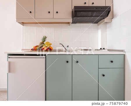 Closeup of white tile splashback on empty wall in simple cabinet module of modern interior kitchen with small fridge, vent, sink, stove, fruits plate 75879740