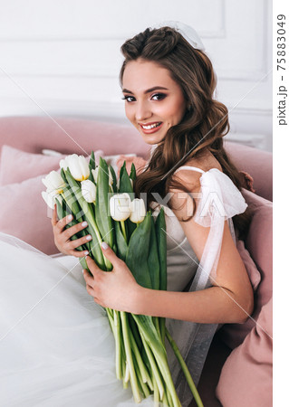 Bride in beautiful dress holding wedding bouquet and sitting on sofa indoors 75883049