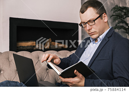 Young successful man in suit and glasses works with notepad and laptop while sitting in spacious home office on couch against the background of fireplace. Success and time management concept. Close-up Young successful man in suit and glasses works with notepad and laptop while sitting in spacious home office on couch against the background of fireplace. Success and time management concept. Close-up 75884114