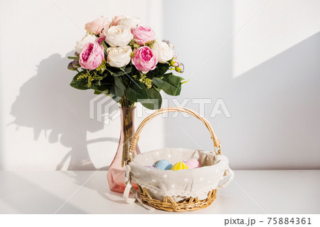 Easter colorful eggs in busket on white wooden table. Bouquet of rose flowers on background. Happy Easter. Springtime mood. 75884361