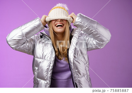 Delighted carefree charming happy young blond woman having fun hiding face pulling hat eyes smiling laughing joyfully enjoying winter sunny day playing siblings standing amused purple background 75885887