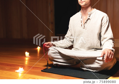 Young Caucasian white male in cotton gray clothes sitting in lotus position in dark meditation room with red rosary in hand. Low key soft focus 75899080