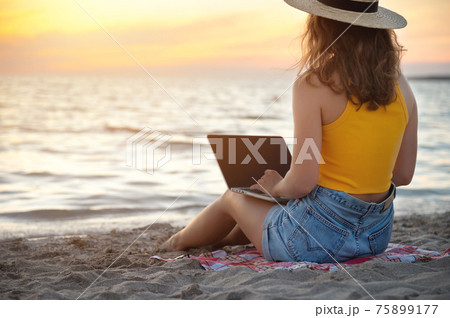 Young Caucasian Woman in shorts and a hat on the beach sitting on the sand browses social networks on the computer in the summer against the background of the sea. Remote work with a laptop 75899177