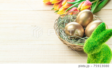 Easter egg. Happy Easter decoration: Golden eggs in basket with spring tulips, white feathers on wooden background. Traditional decoration in sun light. 75913080