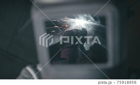 Forge workshop. Smithy. Worker in a welding hood helmet welds a part by electric welding. Sparks are reflected in the protective screen. Forge workshop. Smithy. Worker in a welding hood helmet welds a part by electric welding. Sparks are reflected in the protective screen. 75918956