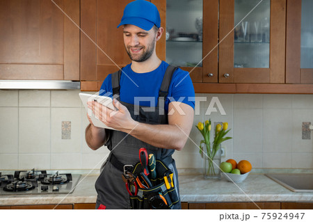 Plumber wearing a tool belt with various tools using tablet during work in kitchen 75924947
