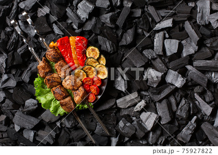 Shish kebab on skewers and grilled sweet pepper, zucchini, cherry, tomato on a round plate. Charcoal background. Shish kebab on skewers and grilled sweet pepper, zucchini, cherry, tomato on a round plate. Charcoal background. 75927822