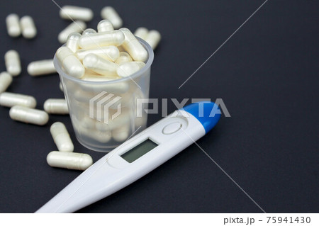 White electronic thermometer and pills in a measuring cup on a black background. Text for space. Selective focus. White electronic thermometer and pills in a measuring cup on a black background. Text for space. Selective focus. 75941430