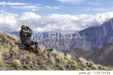 Man in camouflage on the slope is watching through binoculars in the mountains. 75973438
