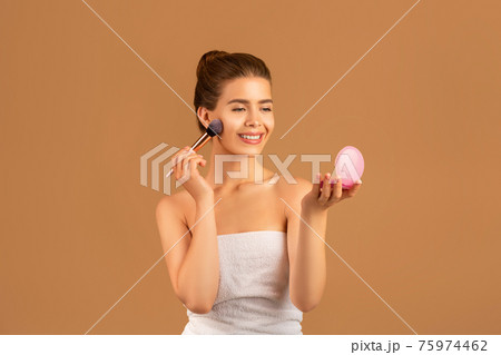 Charming young lady using cosmetic brush to apply decorative makeup, looking in mirror on brown studio background Charming young lady using cosmetic brush to apply decorative makeup, looking in mirror on brown studio background 75974462