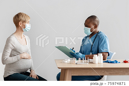 Millennial pregnant woman in protective mask talking to adult african american doctor in clinic sitting at table Millennial pregnant woman in protective mask talking to adult african american doctor in clinic sitting at table 75974871