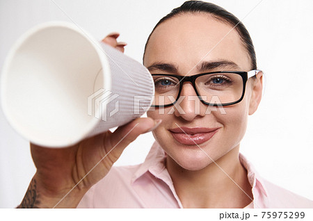 Adorable brunette woman in eyeglasses looks smiling at camera through takeaway paper cup. Closeup, white background, copy space Adorable brunette woman in eyeglasses looks smiling at camera through takeaway paper cup. Closeup, white background, copy space 75975299