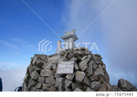 夏空、標高日本第3位の名峰、奥穂高岳山頂にそびえる石造り、切妻屋根の穂高神社嶺宮。標高と山名の看板。 75975495