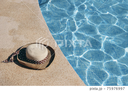 Poolside still life. Straw hat on floor next to resort swimming pool. Glittering crystal clear blue water in sunlight. Summer holiday composition. Swimming, relaxation and travel concept. 75976997