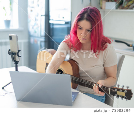 A woman sits in the kitchen during a remote acoustic guitar lesson. A girl learns to play the guitar and watches educational videos on a laptop A woman sits in the kitchen during a remote acoustic guitar lesson. A girl learns to play the guitar and watches educational videos on a laptop 75982294