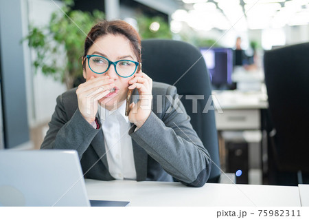 A woman sits in an office at her desk and gossips on the phone. Corporate ethics. Female employee in a suit tells secrets on a smartphone at work. 75982311