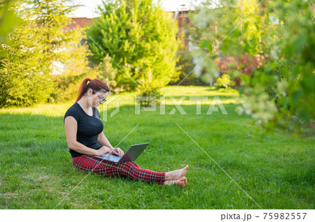 A red-haired woman sits in green grass in a park and is shopping online. The girl maintains a social distance and works outdoors on a laptop. A student is studying remotely on a computer. A red-haired woman sits in green grass in a park and is shopping online. The girl maintains a social distance and works outdoors on a laptop. A student is studying remotely on a computer. 75982557