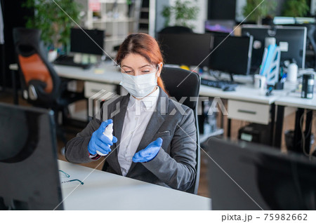 Female manager is sitting in a chair at the workplace. A business woman in a mask is spraying gloves with an antiseptic. Concept of office work during the coronavirus epidemic. Personal hygiene. 75982662