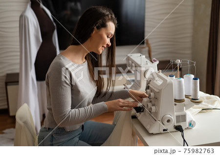 Caucasian woman sews to order at home. Work space of a seamstress in the living room Caucasian woman sews to order at home. Work space of a seamstress in the living room 75982763