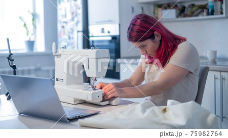 Caucasian woman learning to sew from a video tutorial while sitting in the kitchen. Home hobby for an online lesson Caucasian woman learning to sew from a video tutorial while sitting in the kitchen. Home hobby for an online lesson 75982764