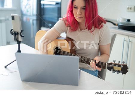 A woman sits in the kitchen during a remote acoustic guitar lesson. A girl learns to play the guitar and watches educational videos on a laptop A woman sits in the kitchen during a remote acoustic guitar lesson. A girl learns to play the guitar and watches educational videos on a laptop 75982814