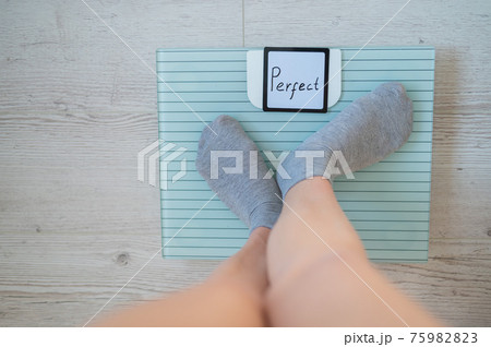Faceless woman measures weight on the floor scales. Top view of women's feet in gray socks on the scales with the word perfect. The inscription on the screen is perfect. There is no excess fat. 75982823