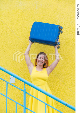 Happy woman in a yellow dress holds a blue suitcase over her head against the background of a yellow wall. Caucasian girl smiles with teeth and happily waits for a trip. Happy woman in a yellow dress holds a blue suitcase over her head against the background of a yellow wall. Caucasian girl smiles with teeth and happily waits for a trip. 75982879