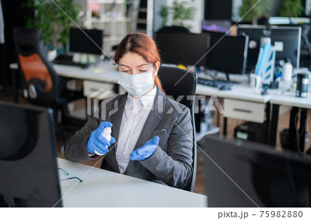 Female manager is sitting in a chair at the workplace. A business woman in a mask is spraying gloves with an antiseptic. Concept of office work during the coronavirus epidemic. Personal hygiene. 75982880